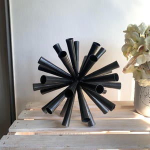 May include: A black metal sculpture shaped like a starburst or sphere, composed of numerous hollow, conical tubes radiating outward from a central point. The object features a dark, matte finish and a symmetrical, multi-directional design.