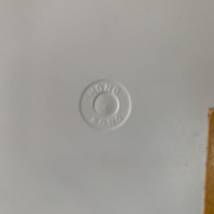 May include: A white surface with a circular, embossed marking in the center. The marking reads "HONG KONG" in a circular pattern. A small, rectangular piece of brown material is visible on the right side.