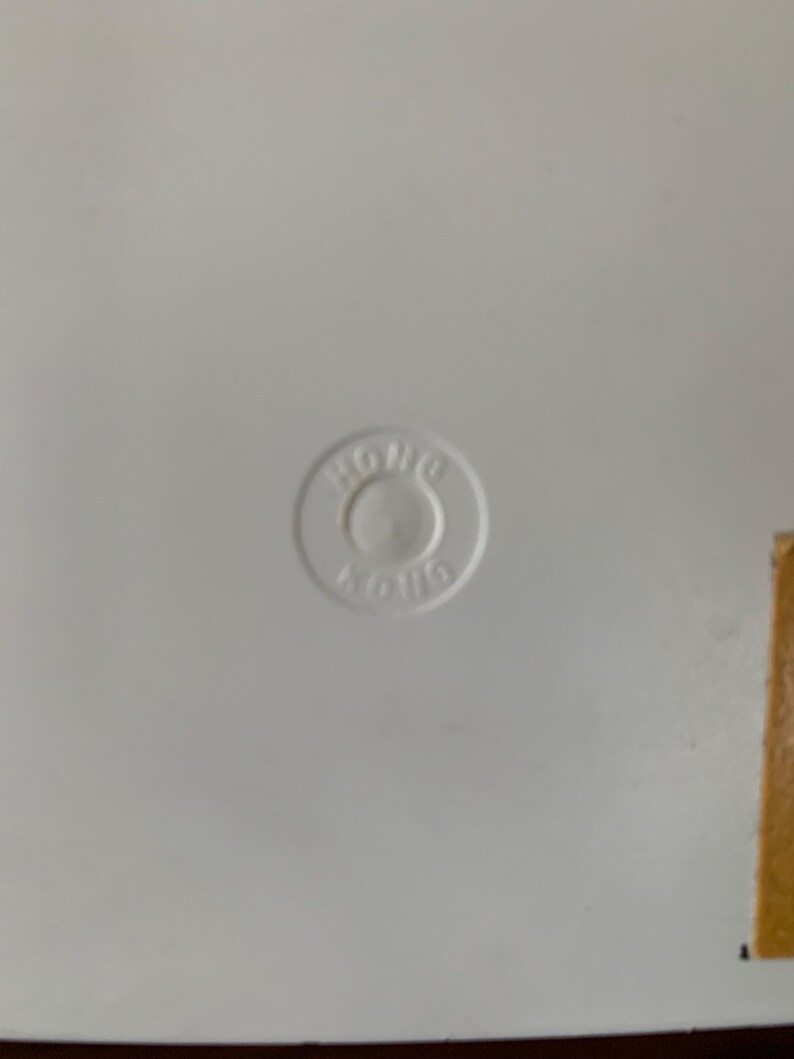 May include: A white surface with a circular, embossed marking in the center. The marking reads "HONG KONG" in a circular pattern. A small, rectangular piece of brown material is visible on the right side.