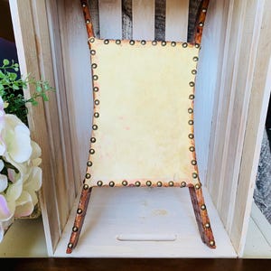 May include: A small, decorative chair with a rectangular seat made of light-colored material. The chair has four wooden legs and a wooden frame. The seat is attached to the frame with small, round, metal rivets. The legs are angled outwards.