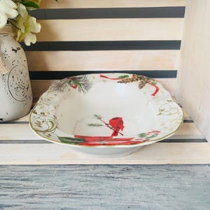 May include: A white ceramic serving bowl with a red cardinal and pine cone design. The bowl has a gold rim and a scalloped edge.