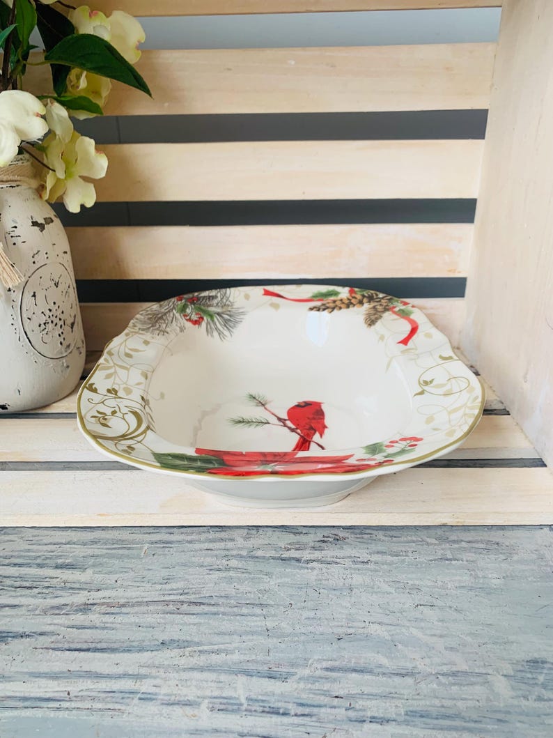 May include: A white ceramic serving bowl with a red cardinal and pine cone design. The bowl has a gold rim and a scalloped edge.