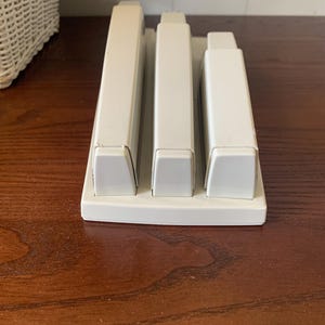May include: A white plastic desk organizer with three rectangular compartments. The organizer sits on a dark wooden surface. The compartments are designed to hold pens, pencils, or other office supplies.