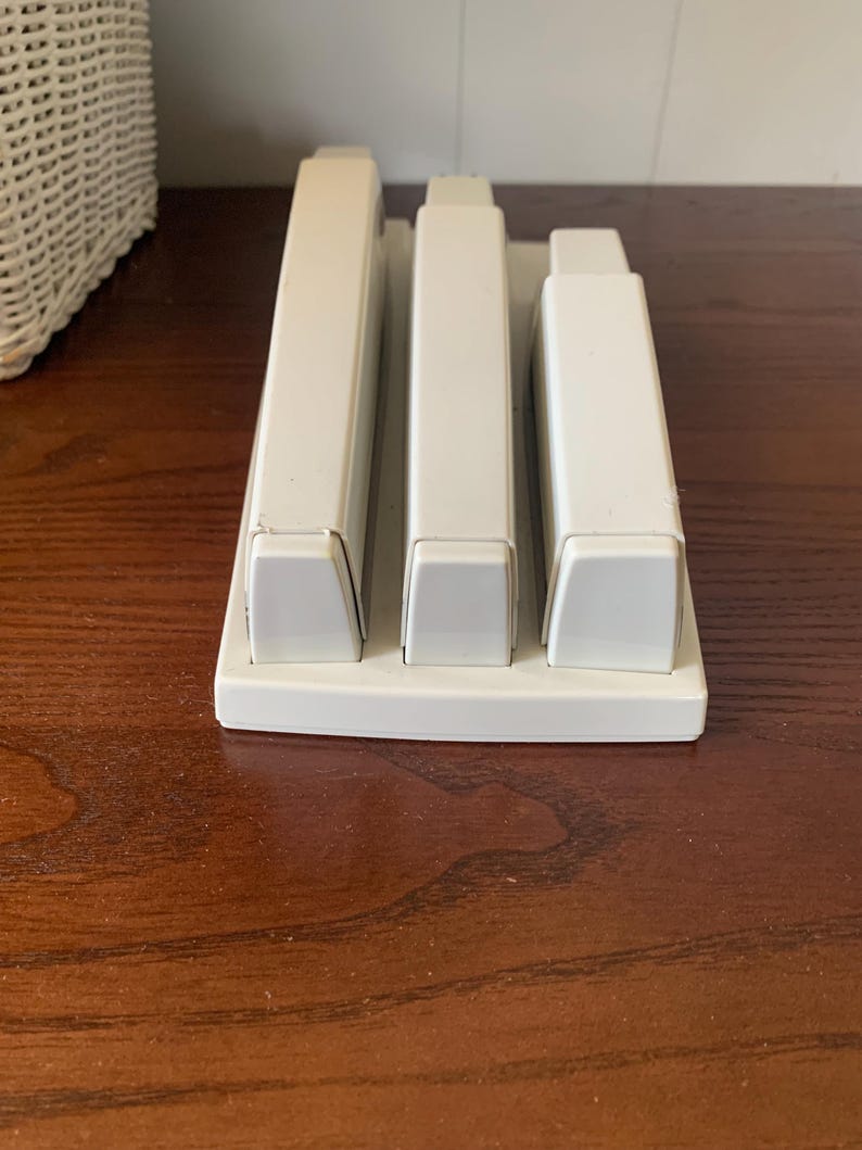 May include: A white plastic desk organizer with three rectangular compartments. The organizer sits on a dark wooden surface. The compartments are designed to hold pens, pencils, or other office supplies.