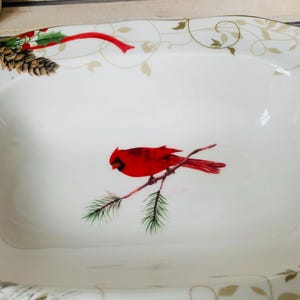 May include: White ceramic serving dish with a red cardinal bird perched on a branch with green pine needles. The dish has a gold and green floral design around the rim.