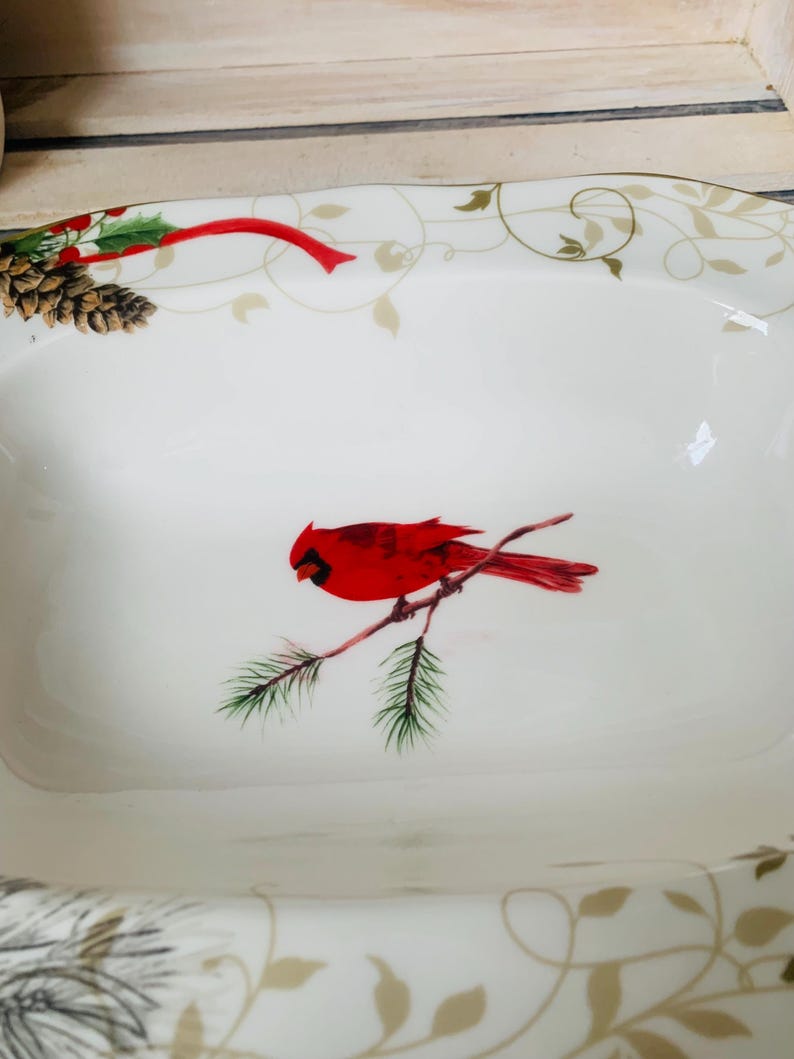 May include: White ceramic serving dish with a red cardinal bird perched on a branch with green pine needles. The dish has a gold and green floral design around the rim.