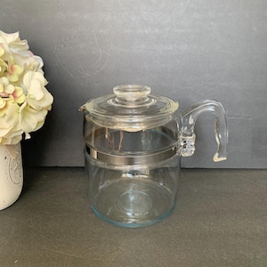 May include: A clear glass coffee pot with a lid and handle. The pot has a silver-coloured band around the middle. The handle is curved and attached to the side of the pot. The pot is on a dark surface.