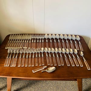 May include: A full set of vintage cutlery arranged on a wooden table. The set includes knives, forks, and spoons with clear acrylic handles and stainless steel heads. The table is brown and has a mid-century modern design.