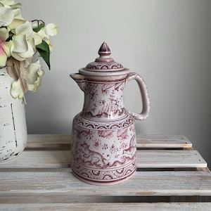 May include: A ceramic pitcher with a lid, featuring a cream-colored base and intricate maroon-colored illustrations of animals, floral patterns, and geometric borders. The pitcher has a curved handle and a pointed lid finial.