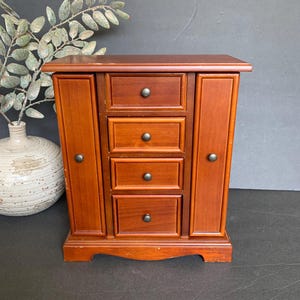 May include: A small, wooden jewelry armoire with a warm, reddish-brown finish. It features three drawers with dark knobs and two side doors. The armoire is set against a dark background, with a decorative vase and foliage visible on the left.