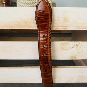 Puede incluir: Un cintur&oacute;n de cuero marr&oacute;n con un patr&oacute;n texturizado similar al de un cocodrilo. El cintur&oacute;n tiene varios agujeros para ajustar la talla y una punta redondeada. El fondo incluye una correa beige tejida y una superficie de madera clara.