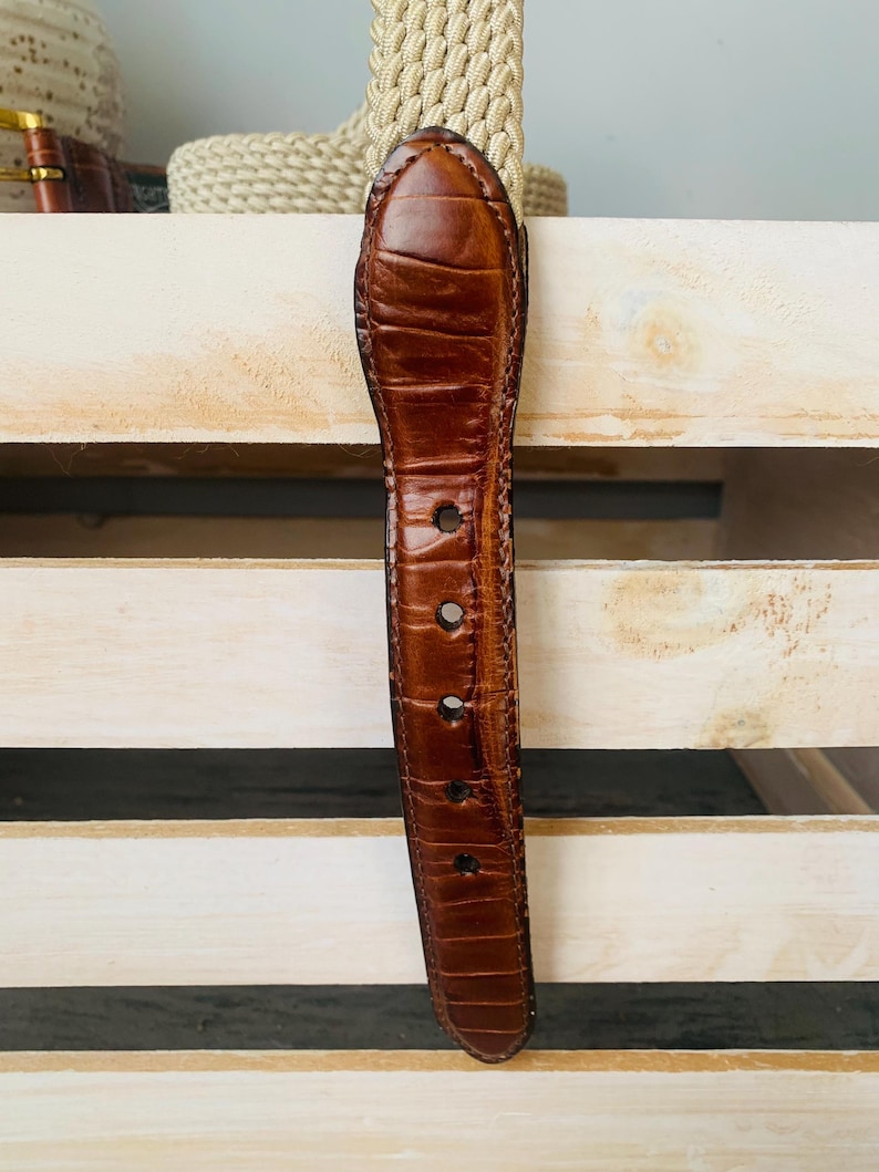 Puede incluir: Un cintur&oacute;n de cuero marr&oacute;n con un patr&oacute;n texturizado similar al de un cocodrilo. El cintur&oacute;n tiene varios agujeros para ajustar la talla y una punta redondeada. El fondo incluye una correa beige tejida y una superficie de madera clara.