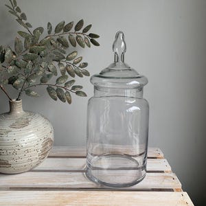 May include: A clear glass apothecary jar with a cylindrical body and a matching glass lid featuring a decorative finial top. The jar is transparent, smooth, and sits upright, showcasing a simple design with a wide opening and a rounded base.