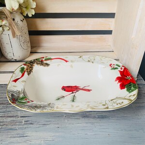May include: White oval serving dish with a gold rim. The dish features a red cardinal bird perched on a branch with green pine needles and red poinsettia flowers.
