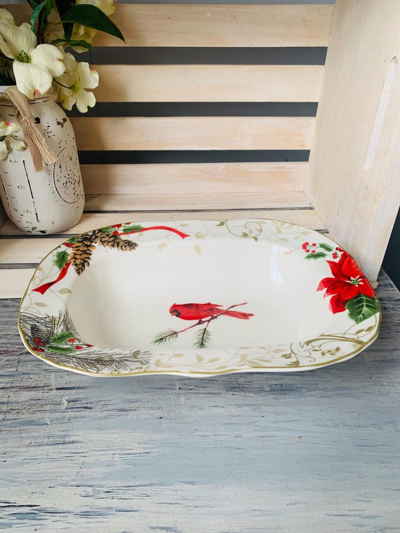 May include: White oval serving dish with a gold rim. The dish features a red cardinal bird perched on a branch with green pine needles and red poinsettia flowers.