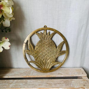May include: A brass pineapple wall hanging. The pineapple is centered within a circular frame, with detailed leaves and a textured surface. The wall hanging is a warm gold colour and has a small hole at the top for hanging.