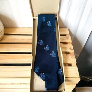 May include: A navy blue tie with a repeating pattern of a flag and the text "1776" in white. The tie is displayed inside a cardboard box with a light brown interior. The tie is made of a textured fabric.