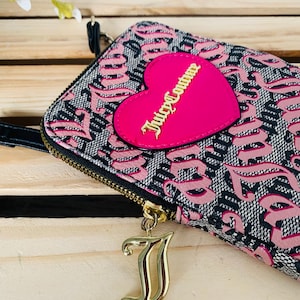 May include: A black and pink wallet with a repeating pattern and a pink heart-shaped patch. The patch has the words "Juicy Couture" in gold lettering. A gold-coloured charm hangs from the zipper.
