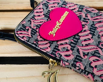 May include: A black and pink wallet with a repeating pattern and a pink heart-shaped patch. The patch has the words "Juicy Couture" in gold lettering. A gold-coloured charm hangs from the zipper.