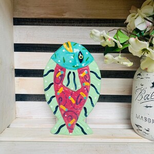 May include: A colorful ceramic fish figurine with a turquoise head, light green body, and a red heart-shaped design. The fish has black dotted lines and is displayed on a white wooden shelf.