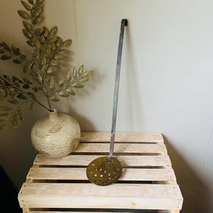 May include: A vintage metal skimmer with a long handle and a round, perforated head. The skimmer is a dull brass colour and sits on a wooden crate. A decorative vase with gold-coloured leaves is in the background.