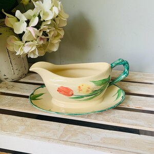 May include: Cream-colored gravy boat and saucer set with a floral tulip design. The boat has a green handle and is paired with a matching saucer. The set is decorated with red and yellow tulips and green leaves.