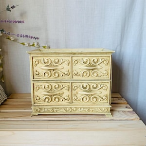 Peut inclure: Une boîte à bijoux de style vintage, de couleur crème, avec quatre tiroirs. La boîte présente des sculptures ornées et tourbillonnantes et de petites poignées incurvées. La boîte est posée sur une surface en bois, avec un fond clair.