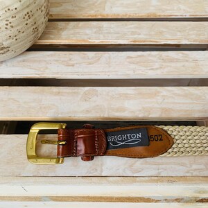 Puede incluir: Un cintur&oacute;n beige tejido con un acento de cuero marr&oacute;n y una hebilla dorada. El cintur&oacute;n tiene una etiqueta que dice "BRIGHTON 502". El cintur&oacute;n se muestra sobre una superficie de madera clara.