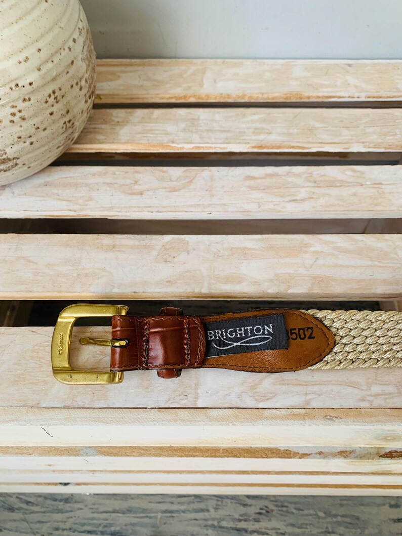 Puede incluir: Un cintur&oacute;n beige tejido con un acento de cuero marr&oacute;n y una hebilla dorada. El cintur&oacute;n tiene una etiqueta que dice "BRIGHTON 502". El cintur&oacute;n se muestra sobre una superficie de madera clara.