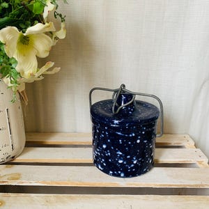 May include: A navy blue ceramic jar with a lid and a metal handle. The jar has a speckled white pattern. The jar is sitting on a wooden crate, with a vase of flowers in the background.