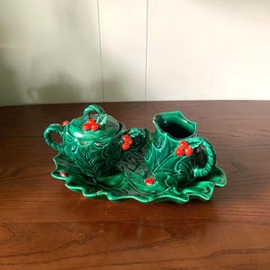 Op de afbeelding: Een groene keramische theeset met een hulstbladontwerp. De set bevat een theepot met deksel, een melkkan en een dienblad. Rode bessenaccenten zijn verspreid over het ontwerp. De set staat op een houten oppervlak.