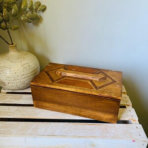 May include: A rectangular wooden box with a decorative handle and geometric design on the lid. The box is made of stained wood with a medium brown colour. The box is sitting on a white wooden crate.