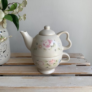 May include: A cream-colored ceramic tea-for-one set consisting of a teapot with a lid and a matching teacup stacked underneath. The set features a hand-painted floral design with soft pink roses and green leaves, and the teapot has a heart-shaped handle.