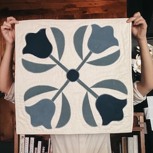 Puede incluir: Un edredón cuadrado de color crema con un diseño floral. Cuatro formas de tulipanes azules estilizados están dispuestas alrededor de un círculo central azul oscuro. El edredón se sostiene, mostrando el diseño.