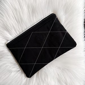 May include: A black quilted pouch with white stitching in a diamond pattern. The pouch is resting on a fluffy white faux fur rug. The background includes a wooden surface and a cowhide rug.