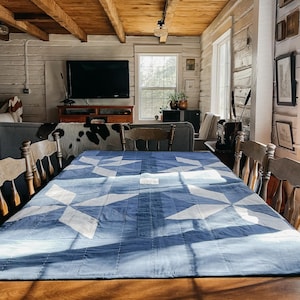 May include: A blue and white star-patterned quilt covers a wooden table, surrounded by wooden chairs. The room features a rustic aesthetic with exposed wooden beams and a cowhide rug.