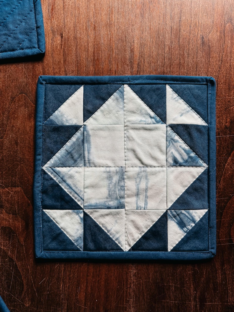 以下が含まれることがあります： 幾何学模様の正方形のキルト鍋つかみ。濃い青色の縁取りと、白と青の三角形と正方形のパターンが中央にあります。生地はタイダイ風です。鍋つかみは木製の表面に置かれています。