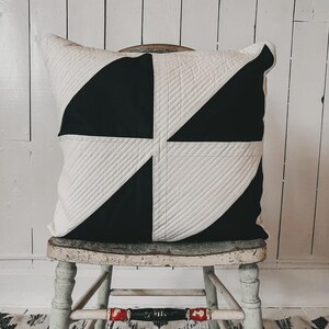 May include: A square decorative pillow with a geometric design. The pillow features a black and white pattern with a quilted texture. The pillow is resting on a distressed wooden chair with a black and white patterned rug in the background.