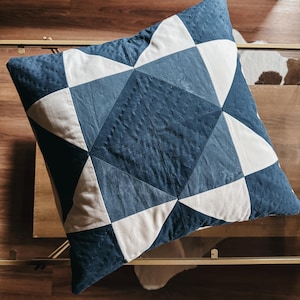 Może przedstawiać: Kwadratowa poduszka dekoracyjna z geometrycznym wzorem patchwork w odcieniach niebieskiego i białego. Poduszka ma pikowaną fakturę i jest umieszczona na stole ze szklanym blatem.