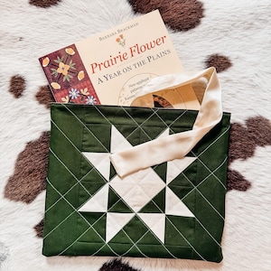 May include: A green tote bag with a white star design, containing a book titled "Prairie Flower: A Year on the Plains." The bag has a cream handle and is on a cowhide rug.