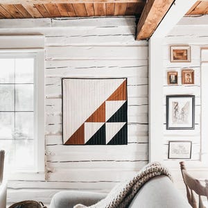 May include: A modern quilt with a geometric design in white, brown, and black hangs on a white-painted log wall. The quilt is square and features triangles and squares. A gray sofa and a woven basket are in the foreground.