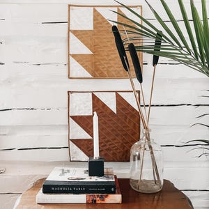 May include: A still-life arrangement featuring two quilted wall hangings with brown and white geometric patterns. A stack of books, a candle, and a glass vase with cattails sit on a wooden table. Green palm fronds add a natural touch.