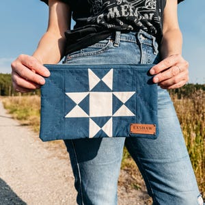Könnte beinhalten: Marineblaue, gesteppte Clutch mit weißem Sternmuster. Die rechteckige Clutch hat ein kleines braunes Lederetikett mit der Aufschrift "EXSHAW". Die Clutch wird von einer Person gehalten, die ein schwarzes T-Shirt und eine blaue Jeans trägt.