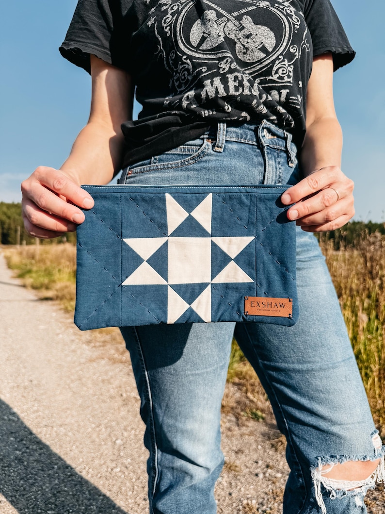 Puede incluir: Bolso de mano acolchado azul marino con un dise&ntilde;o de estrella blanca. El bolso tiene una etiqueta de cuero que dice "EXSHAW". La persona que sostiene el bolso lleva una camiseta negra y vaqueros azules.