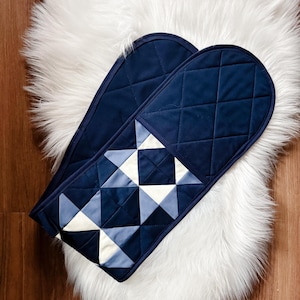 Puede incluir: Dos manoplas de horno azul marino con un diseño geométrico de triángulos blancos y azules. Las manoplas están acolchadas y tienen una textura suave y aterciopelada. Se colocan sobre una alfombra de piel sintética blanca.