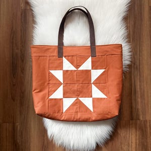May include: An orange tote bag with a white starburst design. The bag has brown handles and is made of quilted fabric. The bag is sitting on a white faux fur rug.