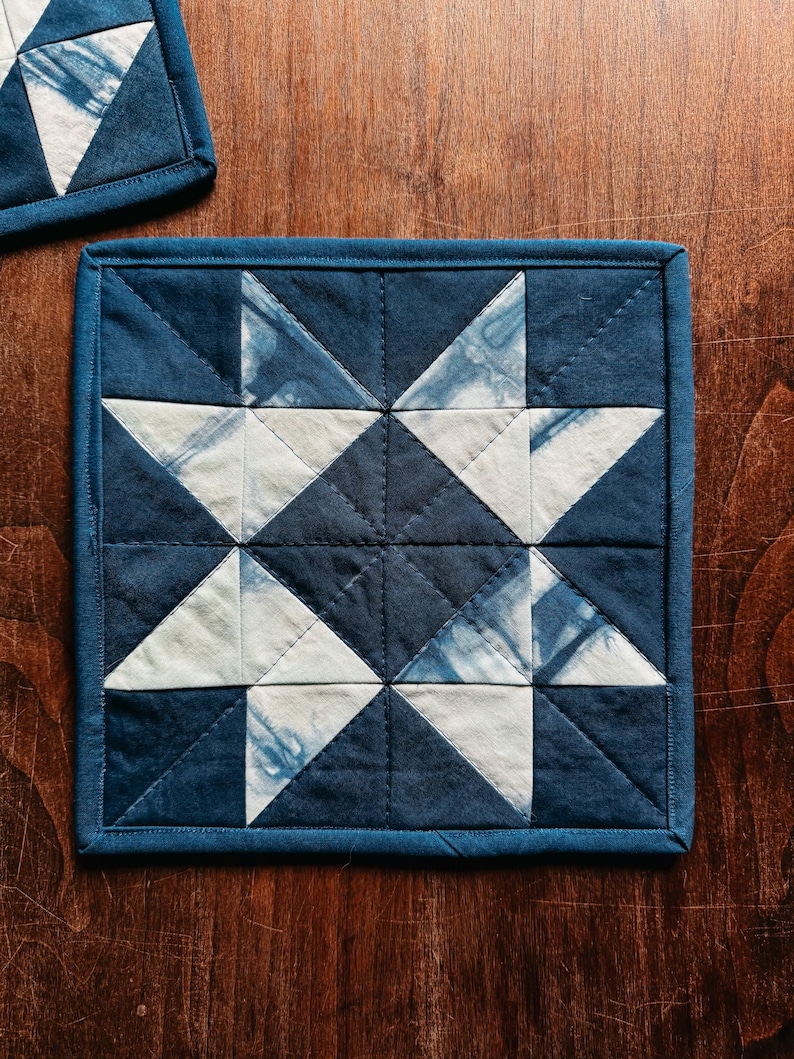 以下が含まれることがあります： 星型のデザインが施された、正方形のキルトコースター。コースターは、ネイビーブルーと白の配色です。デザインは三角形と正方形で構成されています。コースターのサイズは約15cm&times;15cmです。