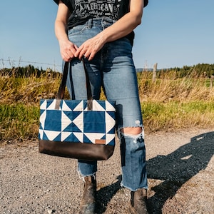 Op de afbeelding: Een draagtas met een geometrisch blauw-wit patroon en een bruine leren basis. De tas heeft bruine leren handvatten en een klein rechthoekig label. De persoon op de foto draagt een zwart t-shirt, een blauwe jeans en bruine laarzen.