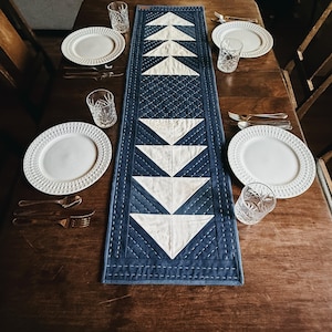 Pode incluir: Uma mesa de jantar de madeira escura para quatro pessoas com pratos brancos, copos de cristal e talheres. Uma toalha de mesa acolchoada azul marinho e branca com um design geométrico corre no centro da mesa. A mesa está posta.