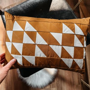 May include: A rectangular decorative pillow with a geometric pattern of alternating brown and white triangles. The pillow is made of fabric and has a quilted design. The pillow is resting on a black chair.